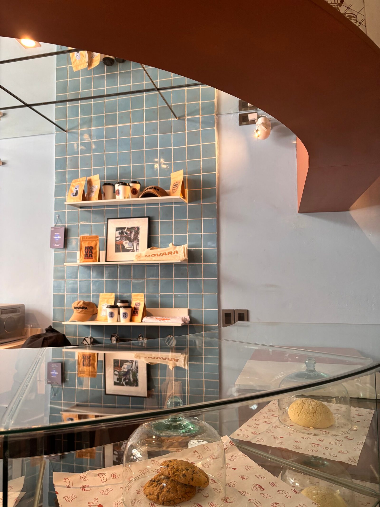 Estante con productos y accesorios de la cafetería Novara en Puebla, con una pared de azulejos azules y una vitrina con pan dulce y galletas.